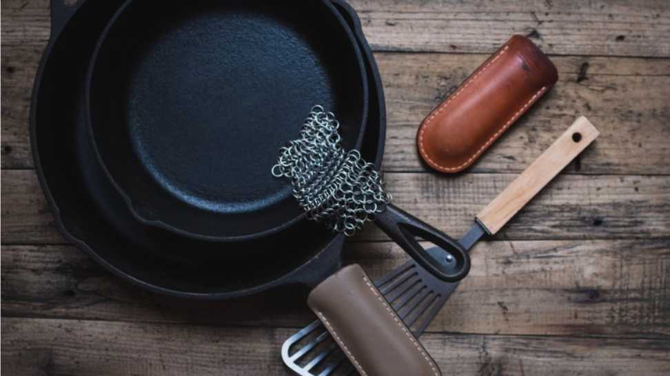 cast iron skillets with a metal spatula leather handle sleeves and a chainmail scrubber on a wooden background