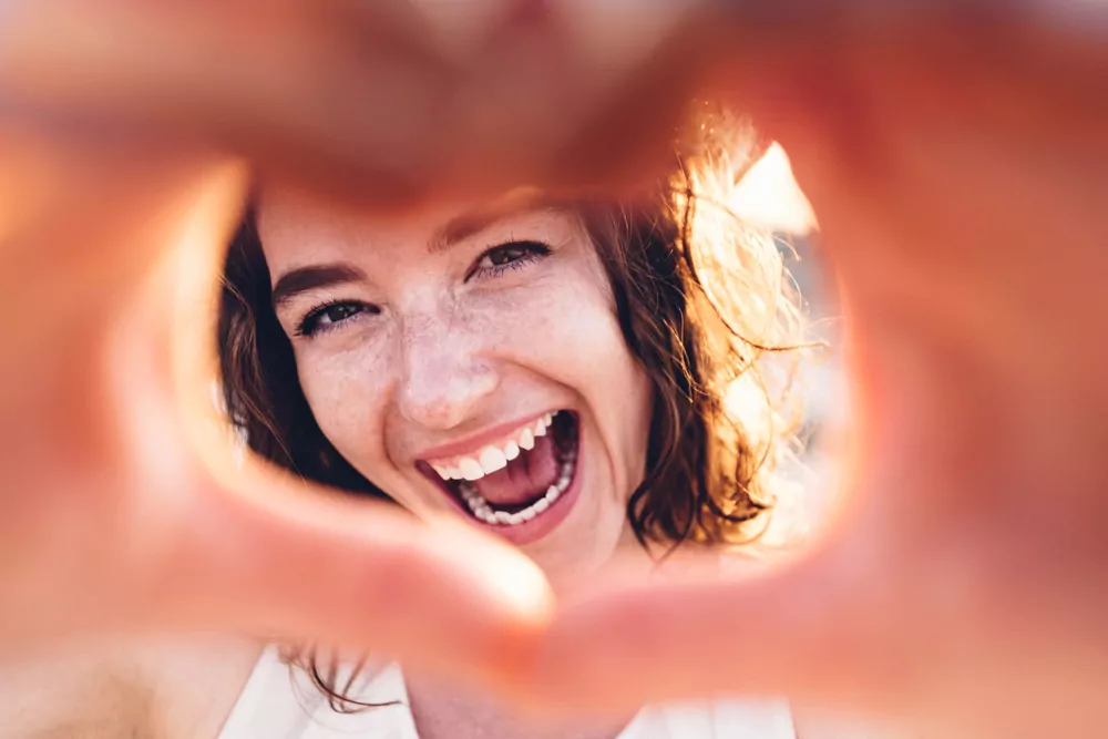 A happy smiling woman making the heart shape with her hands on the camera.