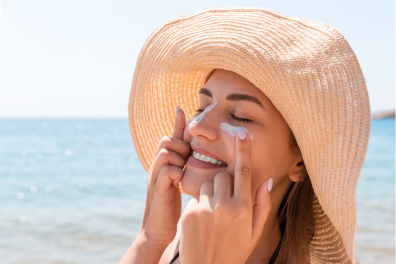 a smiling girl wearing hat and applying sunscreen to her face
