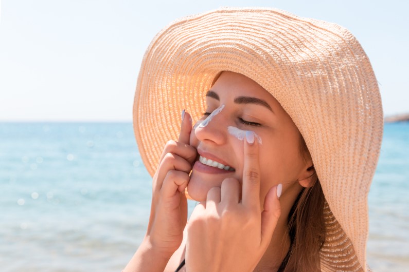 woman in hat is applying sunscreen on her face
