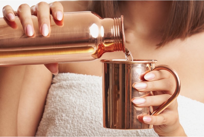 a woman pouring water from a copper bottle into a copper mug