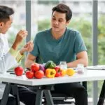 male nutritionist showing and discussing vitamin pills with asian man while sitting at table with healthy food while working in modern hospital