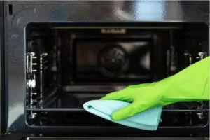 front view of woman in green gloves opening the oven door, wiping dirt at the baking tray inside a microwave oven