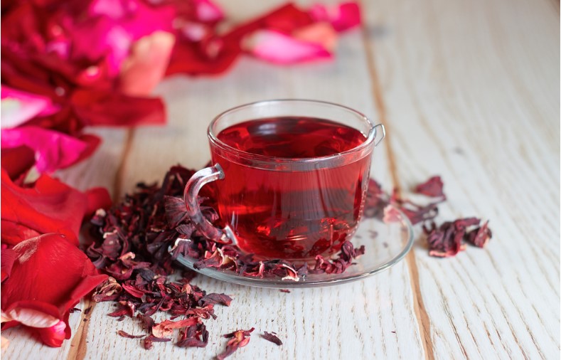 red hot hibiscus tea in a glass mug on a wooden table among rose petals and dry tea custard with metallic heart
