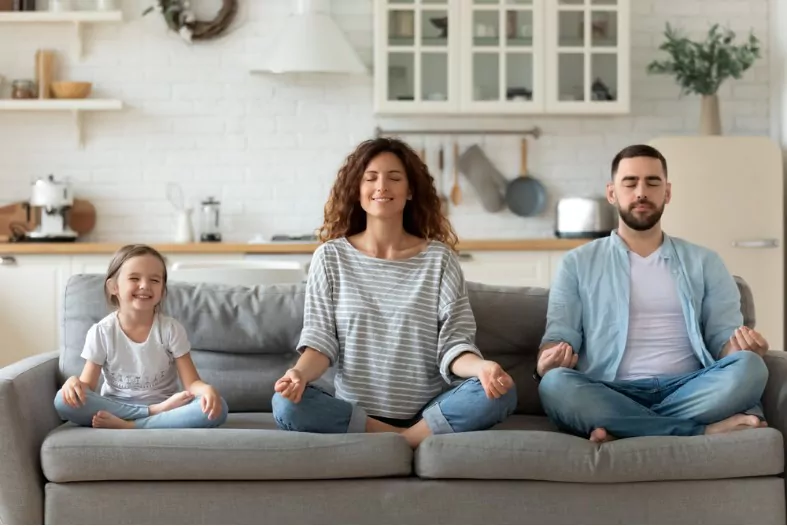 calm young family with little daughter sit on couch practice yoga together