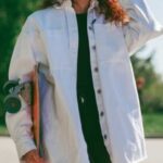a girl in a white oversize shirt holds a skateboard against a backdrop of green trees