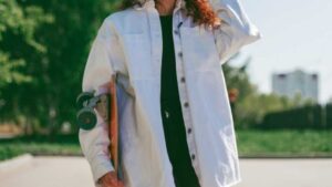 a girl in a white oversize shirt holds a skateboard against a backdrop of green trees